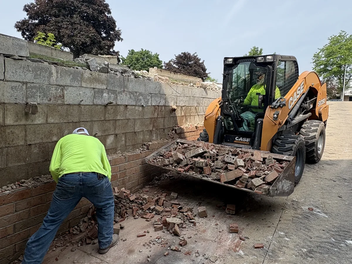 Worker removing brick from a stone wall while a skid steer loads the debris
