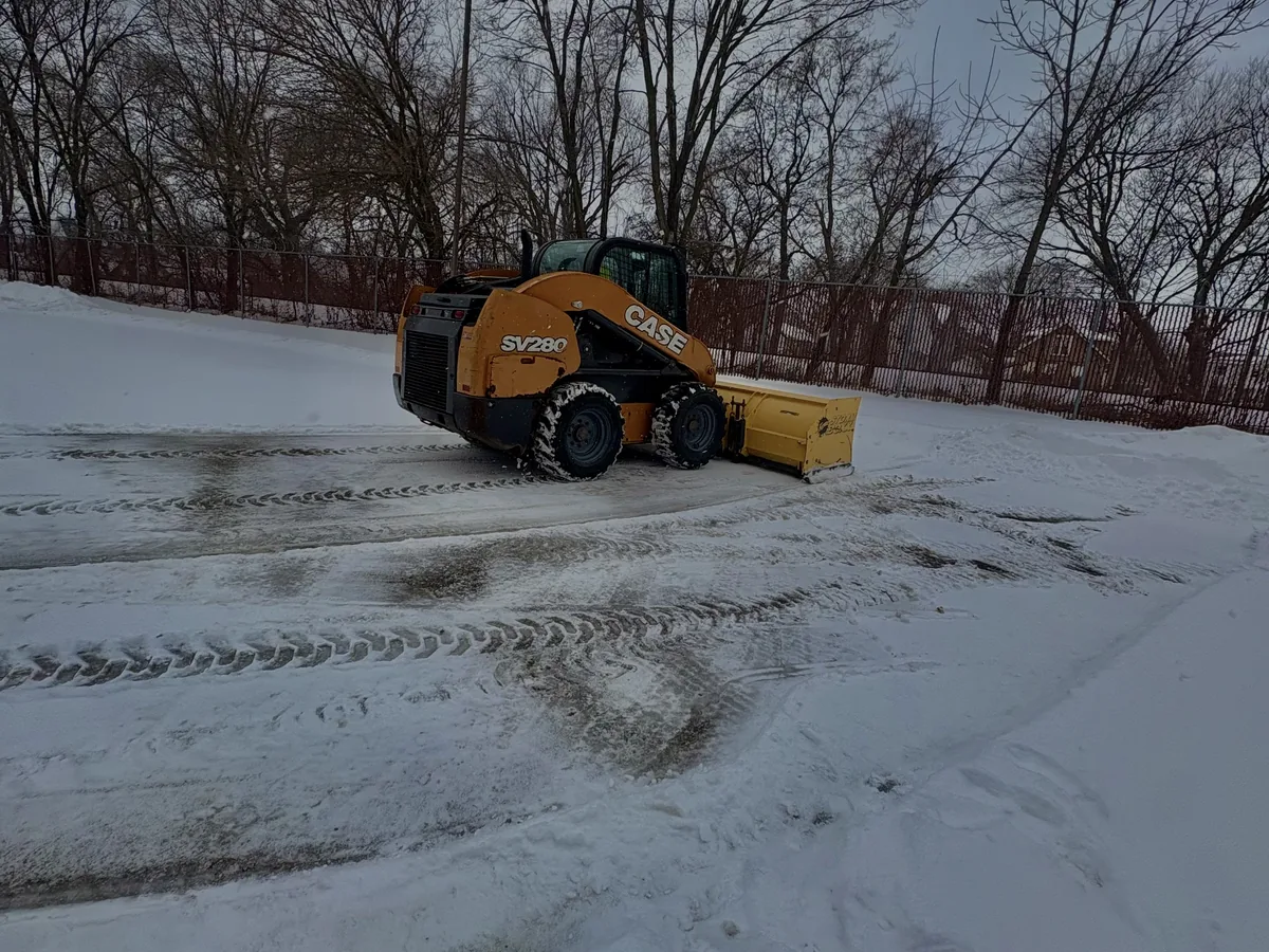 Skid steer with a plow blade clearing snow from a parking lot